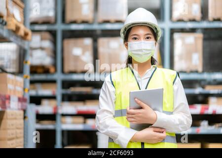 Junge asiatische Frau Auditor oder Trainee-Mitarbeiter trägt Maske während der COVID Pandemie im Lager Versand Industrie arbeiten. Looking up und Kontrollen Stockfoto