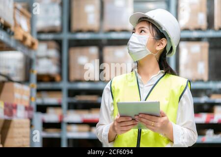 Junge asiatische Frau Auditor oder Trainee-Mitarbeiter trägt Maske während der COVID Pandemie im Lager Versand Industrie arbeiten. Looking up und Kontrollen Stockfoto