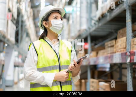 Junge asiatische Frau Auditor oder Trainee-Mitarbeiter trägt Maske während der COVID Pandemie im Lager Versand Industrie arbeiten. Looking up und Kontrollen Stockfoto