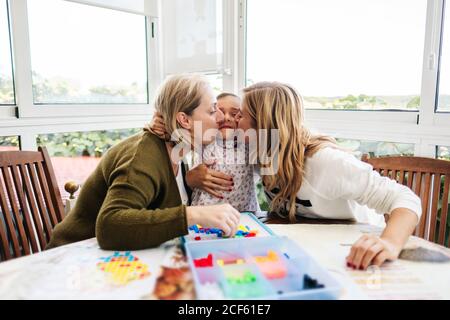 Frau mittleren Alters mit kleinen Mädchen und Erwachsene Tochter Spaß haben und Brettspiel Umarmung und küssen, während sitzen am runden Tisch auf der Terrasse Stockfoto