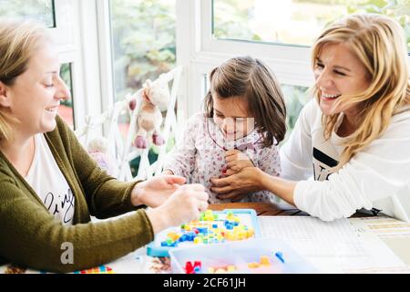 Frau mittleren Alters mit kleinen Mädchen und Erwachsene Tochter Spaß haben und Brettspiel spielen, während lachen und kitzeln sich am Tisch Stockfoto