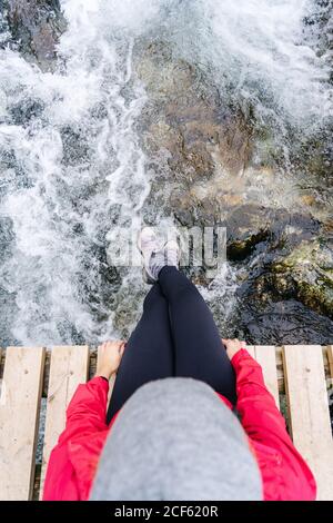 Von oben Ansicht der unkenntlich entspannten touristischen Frau in roter Jacke mit Rucksack sitzen und baumelnde Beine auf Holzbrücke im Dorf in den Gipfeln Europas, Asturien, Spanien Stockfoto
