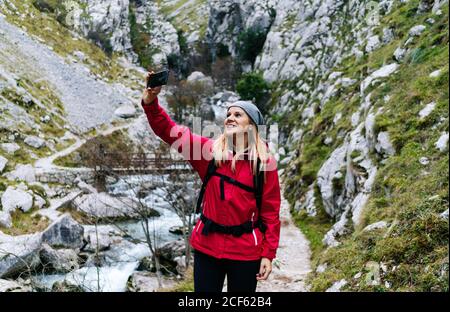 Frau Tourist in roter Jacke mit Rucksack Selfie auf dem Handy auf dem Hintergrund der grünen felsigen Berge im Dorf in den Gipfeln Europas, Asturien, Spanien Stockfoto