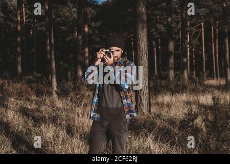 Bärtiger Mann im lässigen Outfit, der mit der Kamera fotografiert Stehend umgeben von immergrünen Bäumen und getrocknetem Gras auf Kälte Sonniger Tag Stockfoto
