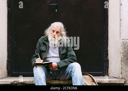 Nachdenkliche ältere männliche Touristen in Freizeitkleidung sitzen mit Rucksack Auf Steinschritt des schäbigen Gebäudes und Schreiben Gedanken über Urlaub im Notebook Stockfoto