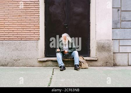 Nachdenkliche ältere männliche Touristen in Freizeitkleidung sitzen mit Rucksack Auf Steinschritt des schäbigen Gebäudes und Schreiben Gedanken über Urlaub im Notebook Stockfoto