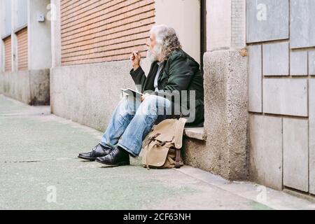 Seitenansicht eines nachdenklichen älteren männlichen Touristen in legerer Kleidung Sitzen mit Rucksack auf Stein Schritt von schäbigen Gebäude und Schreiben von Gedanken über Urlaub in Notebook Stockfoto