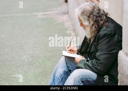 Seitenansicht eines nachdenklichen älteren männlichen Touristen in legerer Kleidung Sitzen mit Rucksack auf Stein Schritt von schäbigen Gebäude und Schreiben von Gedanken über Urlaub in Notebook Stockfoto