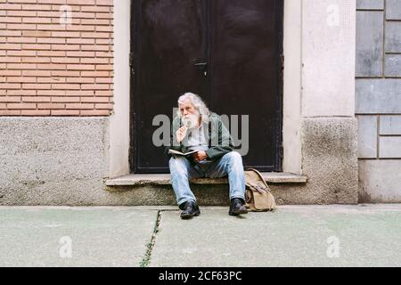 Nachdenkliche ältere männliche Touristen in Freizeitkleidung sitzen mit Rucksack Auf Steinschritt des schäbigen Gebäudes und Schreiben Gedanken über Urlaub im Notebook Stockfoto