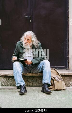 Nachdenkliche ältere männliche Touristen in Freizeitkleidung sitzen mit Rucksack Auf Steinschritt des schäbigen Gebäudes und Schreiben Gedanken über Urlaub im Notebook Stockfoto