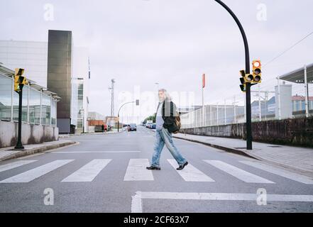 Seitenansicht von reifen männlichen Reisenden in legerer Kleidung zu Fuß Mitten in einer leeren Fußgängerüberfahrt, während man urbane Gebäude erkundet Stockfoto