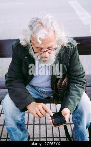 Von oben konzentrierter graubärtiger Mann in lässiger Kleidung tragen Brillen und Messaging auf dem Handy, während Sie auf der Straße ruhen Bank Stockfoto