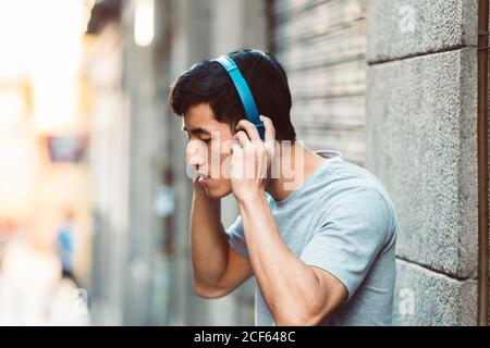 Fröhlicher zwangloser ethnischer asiatischer Mann in Kopfhörern, der Musik hört Während Sie auf der sonnigen Stadtstraße stehen Stockfoto