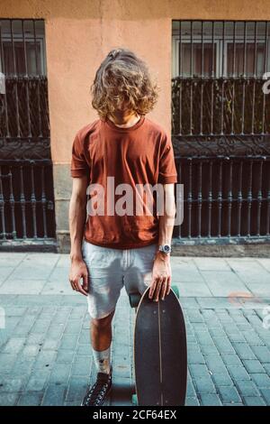 Schöner sportlicher Mann hält Longboard, während er das Gesicht mit bedeckt Haare auf Stadt Sommer Stadt Straße Stockfoto