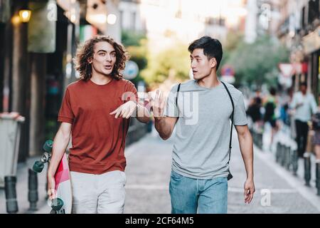 Sorgenfrei interessierte multiethnische Männer in lässiger Kleidung mit Longboard-Gestik Und reden beim Bummeln entlang der Stadtstraße Stockfoto