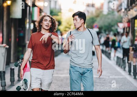 Sorgenfrei interessierte multiethnische Männer in lässiger Kleidung mit Longboard-Gestik Und reden beim Bummeln entlang der Stadtstraße Stockfoto