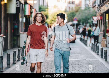 Sorgenfrei interessierte multiethnische Männer in lässiger Kleidung mit Longboard-Gestik Und reden beim Bummeln entlang der Stadtstraße Stockfoto