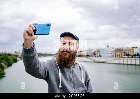 Content männliche Reisende in lässiger Kleidung lächeln und Selfie machen Und Friedenszeichen auf dem Handy, während man auf schaukelte Zaun des Kais Stockfoto