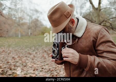 Nicht erkennbare Seitenansicht des erwachsenen Mannes mit Retro-Fotokamera Stehen auf trockenen Blättern im ruhigen Herbstpark im Herbst Stockfoto