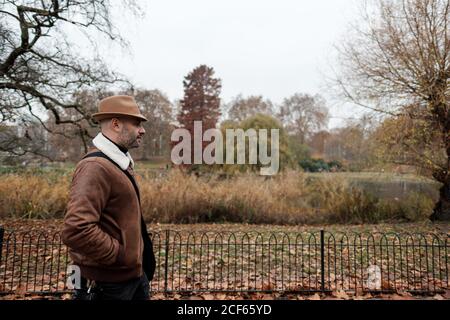 Seitenansicht eines erwachsenen Mannes in legerem Outfit, der die Hände hält In Taschen zu Fuß in der Nähe von Zaun in ruhigen Herbst Park in London Stockfoto