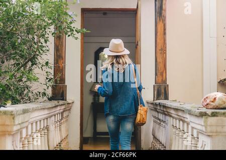 Rückansicht einer jungen Frau in Denim und Hut, die die hölzerne Glastür eines authentischen Gebäudes öffnet Stockfoto