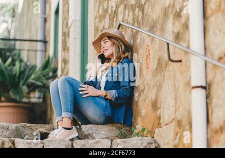 Seitenansicht einer jungen lächelnden Frau, die auf einem Mobiltelefon sprach und draußen auf einer Steintreppe saß Stockfoto