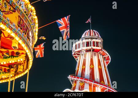 Von unten hell erleuchtete Fahrten mit Fahnen gegen dunklen Nachthimmel auf dem Messegelände in London, Großbritannien Stockfoto