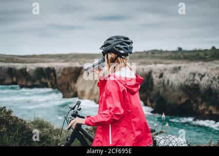 Seitenansicht der Frau auf dem Fahrrad in Helm und roter Jacke Trinkwasser und genießen malerische Landschaft Stockfoto