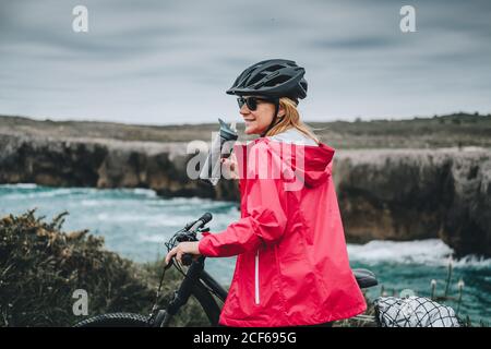 Seitenansicht der Frau auf dem Fahrrad in Helm und roter Jacke Trinkwasser und genießen malerische Landschaft Stockfoto