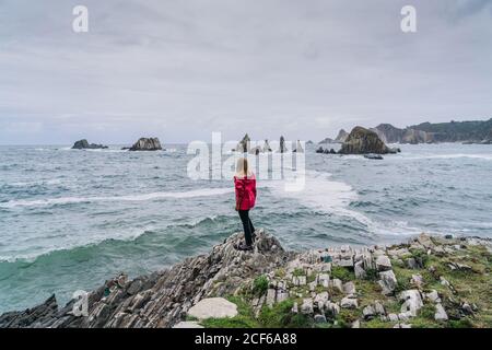 Rückansicht einer Frau in roter Jacke, die auf einem großen Stein am felsigen Meer steht und am bewölkten Tag wellige, schaumig wäschende Wasserspitzen beobachtet Stockfoto