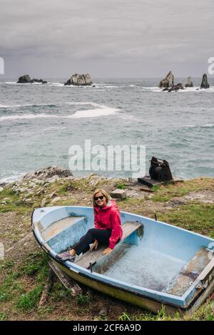 Von oben Frau in roter Jacke im schäbigen blauen Boot am Meer umgeben von welligem Schaum Wasser waschen Steine und Gipfel Stockfoto