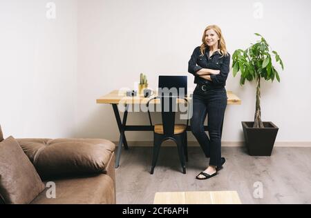 Die schöne blonde kaukasische Frau arbeitet aus ihrem Wohnzimmer mit ihrem Laptop auf einem Holzschreibtisch. Sie trägt schwarze Freizeitkleidung. Stockfoto