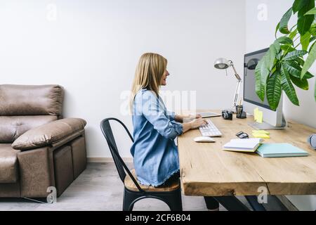 Seitenansicht einer blonden kaukasischen Frau in legerer Kleidung, die auf einem Holzschreibtisch sitzt und an einem Computer in einem modernen Heimbüro arbeitet Stockfoto