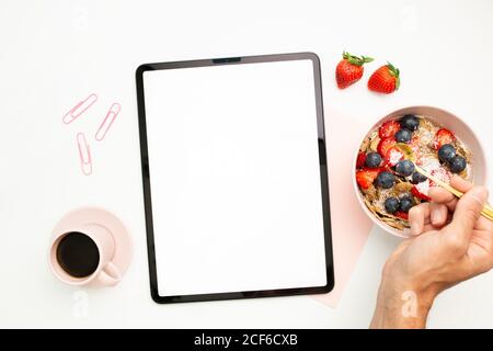 Draufsicht auf unkenntlich Männchen mit Teelöffel in Schüssel mit Frisches Obst und Beeren sitzen am weißen Tisch mit Tasse Von Kaffee und Tablet mit leerem Bildschirm Stockfoto