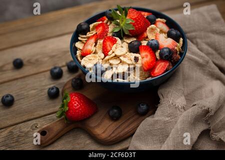 Von oben köstliche Frühstückschale mit Maisflocken mit Erdbeeren Und Heidelbeeren auf Schneidebrett gelegt und mit Leinen verziert Tuch und Beeren rund um Gericht auf Holz Hintergrund Stockfoto