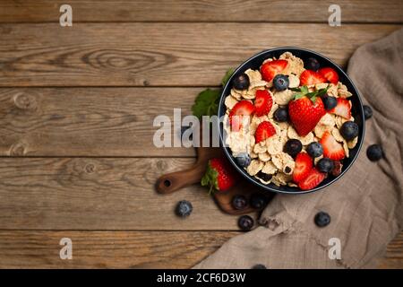 Blick von oben auf das leckere Frühstück Schüssel mit Cornflakes mit Erdbeeren und Heidelbeeren auf Schneidebrett gelegt und mit verziert Leinentuch und Beeren rund um Gericht auf Holzhintergrund Stockfoto