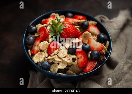 Blick von oben auf das leckere Frühstück Schüssel mit Cornflakes mit Erdbeeren und Heidelbeeren auf Schneidebrett gelegt und mit verziert Leinentuch und Beeren rund um Gericht auf Holzhintergrund Stockfoto