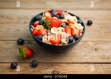 Blick von oben auf das leckere Frühstück Schüssel mit Cornflakes mit Erdbeeren und Heidelbeeren auf Schneidebrett gelegt und mit verziert Leinentuch und Beeren rund um Gericht auf Holzhintergrund Stockfoto