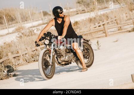 Entzückter Mann in Helm und Sonnenbrille auf dem Motorrad sitzen Feld und wegschauen Stockfoto