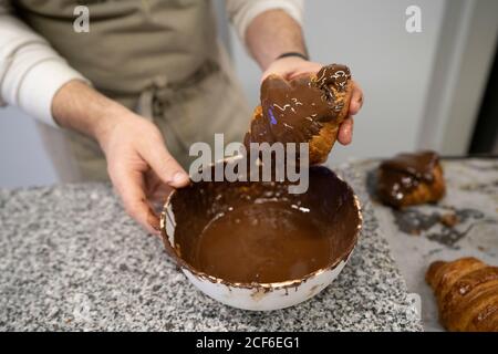 Von oben Ernte Mann in Schürze Dipping knusprige Croissants in Schüssel mit köstlicher Schokoladencreme Stockfoto