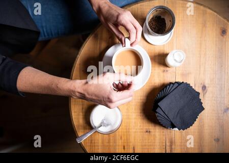 Oben die Hände des Weibchens, das am hölzernen Tisch sitzt Und heißen Tee mit Milch und Zucker rühren Stockfoto