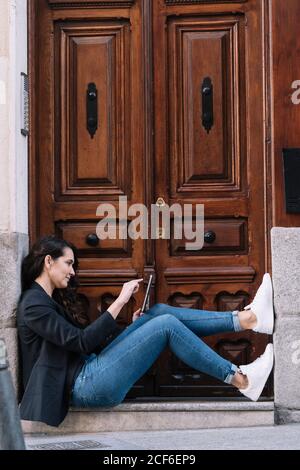 Seitenansicht der schönen Frau mit digitalen Tablet beim Entspannen auf der Schwelle der alten Holztür Stockfoto
