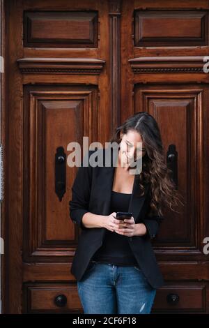 Schöne, fröhliche Frau, die mit dem Mobiltelefon telefoniert und sich entspannt an der alten Holztür lehnt Stockfoto