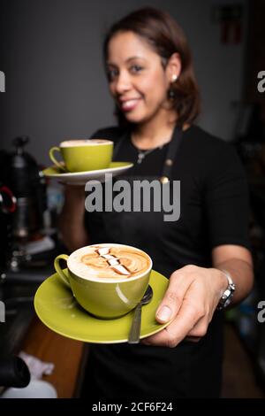 Lächelnde Barista, die grüne Kaffeetassen mit Latte-Art gibt Und Blick auf die Kamera im Café Stockfoto