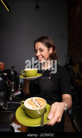 Lächelnde Barista, die grüne Kaffeetassen mit Latte-Art gibt Und Blick auf die Kamera im Café Stockfoto