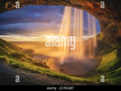 Majestätischer Wasserfall befindet sich am Eingang der grasbewachsenen Steinhöhle gegen wolkiger Sonnenuntergang am Meer in Island Stockfoto