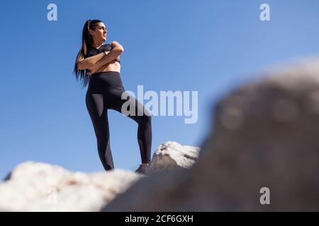 Von unten junge Brünette Frau in Sportkleidung mit gekreuzten Händen posiert auf Felsen in sonnigen Tag Stockfoto