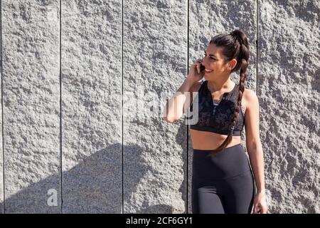 Junge Brünette glückliche Frau in Sportkleidung im Gespräch auf Handy in der Nähe Steinmauer an sonnigen Tag Stockfoto
