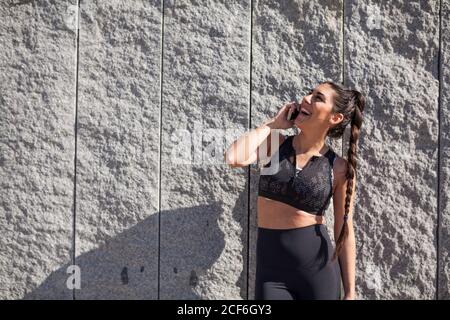 Schöne und brünette Frau, die sich an einer Granitwand lehnt, während sie mit Freunden telefoniert und lacht Stockfoto