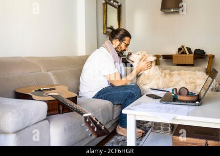 Seitenansicht des glücklichen bärtigen Mannes in legerer Kleidung sitzend Auf der Couch und lächelnd beim Kuscheln mit dem großen Golden Retriever Im modernen Wohnzimmer Stockfoto